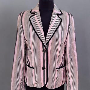 Puede incluir: Blazer a rayas rosas y negras con ribete negro y dos botones negros. El blazer se exhibe en un maniquí. La chaqueta tiene solapas clásicas y dos bolsillos delanteros. El fondo es gris liso.