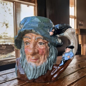 Puede incluir: Una taza de cerámica con un diseño detallado y pintado a mano de un anciano con barba larga y sombrero azul. Un pájaro negro está posado en el asa, con una pequeña figura en el asa. La taza es marrón, azul y beige.