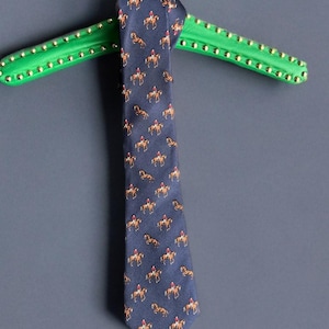 Puede incluir: Una corbata de seda azul oscuro con un patrón repetido de caballos y jinetes marrones. La corbata está colgada de una percha verde con tachuelas.