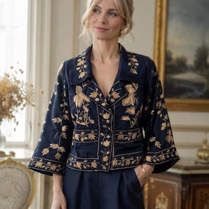 Op de afbeelding: Een marineblauw jasje met een reverskraag en driekwartmouwen, versierd met goudkleurig bloemenborduursel op de voorzijde en de mouwen. Het jasje heeft een knoopsluiting aan de voorzijde en wordt gecombineerd met een bijpassende marineblauwe broek.