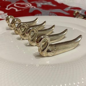 May include: A collection of silver swan-shaped napkin rings arranged on a white plate. The napkin rings are detailed with curved necks and sleek bodies. The plate has a textured rim, and the background features a red and white patterned fabric.