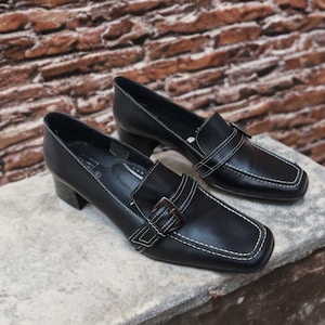 Puede incluir: Un par de mocasines de cuero negro con puntera cuadrada y tacón pequeño. Los zapatos tienen costuras blancas y una correa decorativa con hebilla. Los mocasines se exhiben sobre una superficie de piedra.
