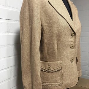 May include: A tan blazer featuring a textured weave, a notched lapel, and three front buttons. The garment includes two front pockets with buttoned flaps and frayed edges, along with matching frayed detailing on the sleeve cuffs and hemline.
