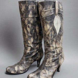 Könnte beinhalten: Ein Paar hohe, abgenutzte Lederstiefel in Braun- und Beigetönen. Die Stiefel haben eine spitze Zehenpartie, einen hohen Absatz und ein dekoratives blattförmiges Detail. Die Stiefel werden vor grauem Hintergrund präsentiert.