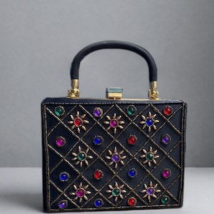 Puede incluir: Un bolso de mano rectangular negro con un patrón de diamantes acolchado delineado con hilo dorado. La superficie está adornada con joyas circulares multicolores y detalles dorados en forma de estrella. Tiene un asa superior negra y un cierre.
