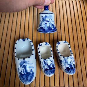 Puede incluir: Colección de zuecos holandeses y una campana de cerámica azul y blanca. Los zuecos son de tres tamaños, y la campana está sostenida por una mano. Los artículos están decorados con un diseño de molino de viento y barco.