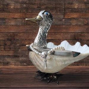 Puede incluir: Un plato decorativo plateado con forma de pato con un collar de perlas y un cierre plateado. El pato tiene ojos azules y un pico dorado. El plato está hecho de una concha y se asienta sobre una base oscura y ornamentada.