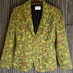 Puede incluir: Una blazer verde con estampado de cachemira y forro negro. La blazer tiene cierre de botón y dos bolsillos delanteros. El estampado de cachemira incluye tonos de verde, marrón y rojo. La etiqueta dice "Vincenzo".