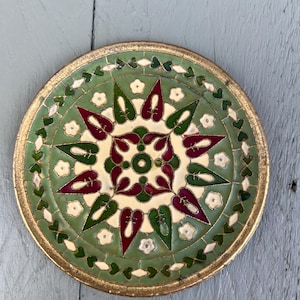 Puede incluir: Un plato decorativo y redondo con borde dorado. El plato presenta un diseño central de formas burdeos y verdes sobre fondo crema, rodeado de un borde verde con detalles florales blancos. El plato tiene unos 20 cm de diámetro.