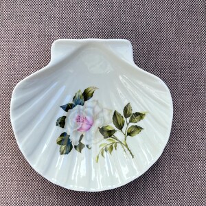 Puede incluir: Un plato de porcelana blanca con forma de concha de vieira, que presenta un diseño pintado de una rosa blanca con un centro rosa suave y hojas verdes. El plato tiene un acabado brillante y una textura acanalada que imita la forma natural.
