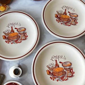 Puede incluir: Platos de cerámica color crema con borde marrón y la palabra "Crêpes" en cursiva. Cada plato presenta una colorida ilustración de crepes, huevos y otros elementos de desayuno. Una adición encantadora a cualquier cocina.