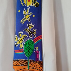 Puede incluir: Una corbata de seda azul con un diseño colorido y caprichoso. La corbata presenta pájaros amarillos, un arcoíris, un árbol verde y un edificio rosa sobre un fondo azul. La punta de la corbata es naranja con líneas negras. La firma del artista es visible.