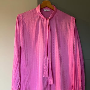 Puede incluir: Una blusa de seda rosa con cuello de lazo y una pequeña etiqueta blanca que dice "CELINE" en el cuello.
