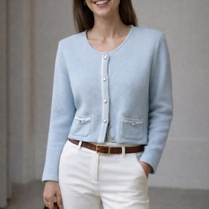 Puede incluir: Un cárdigan azul claro con botones dorados y ribete blanco, combinado con pantalones blancos y un cinturón marrón. El cárdigan tiene dos bolsillos delanteros. Se sostiene un bolso marrón.