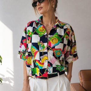 Puede incluir: Una camisa de manga corta con estampado de banderas y medallones de colores. La camisa presenta un patrón de cuadros blancos y negros y se combina con pantalones blancos y un cinturón negro. La modelo lleva gafas de sol y joyas doradas.