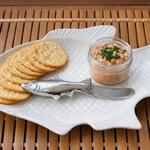 Puede incluir: Una fuente blanca con forma de pez con galletas saladas, un tarro de salsa y un cuchillo de mantequilla con forma de pez. La salsa está cubierta con hierbas verdes. La fuente está sobre una esterilla de bambú.