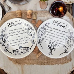 Puede incluir: Dos platos de cerámica blanca con texto e ilustraciones en negro. Un plato presenta un pez, el otro una langosta. Ambos platos tienen un diseño de hojas en el borde. También se ven corchos y pequeñas plantas en macetas.