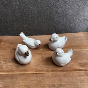 Puede incluir: Cuatro figuras de pájaros de cerámica blanca con picos dorados. Dos pájaros están erguidos y dos en reposo. Los pájaros tienen marcas de plumas detalladas y están dispuestos sobre una superficie de madera.