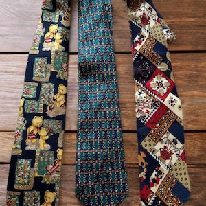 Puede incluir: Tres corbatas estampadas se muestran sobre una superficie de madera. Una corbata presenta un diseño de osos de peluche, otra un patrón geométrico y la tercera un mosaico de elementos florales y geométricos. Las corbatas son en tonos azules, verdes, rojos y beige, ideales para ocasiones formales.