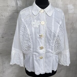 Puede incluir: Blusa blanca abotonada con bordados y detalles de encaje. Presenta un cuello redondo, botones decorativos y mangas tres cuartos. La blusa tiene una cintura entallada y un dobladillo festoneado.