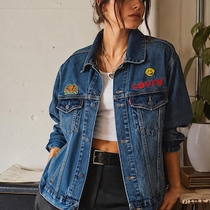 Puede incluir: Una chaqueta de mezclilla azul oscuro con el logotipo de Levi's bordado en rojo en el pecho izquierdo, un parche de cara sonriente amarilla sobre el logotipo y un parche de oso marrón en el pecho derecho. La chaqueta tiene cuello, botones y bolsillos.