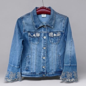 May include: A light blue denim jacket with a classic collar, button-down front, and two chest pockets. The jacket features intricate white floral embroidery on the pockets and cuffs. The cuffs have a flared design with matching white lace detailing. The jacket is hanging on a red hanger.