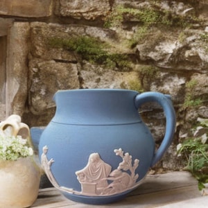 Puede incluir: Jarra de cerámica azul claro con asa y figuras en relieve blancas. La jarra está colocada contra una pared de piedra y un pequeño jarrón con flores blancas.