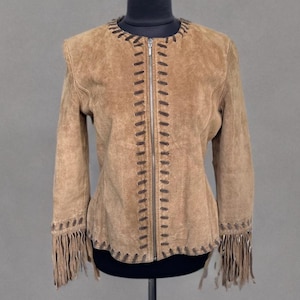 Puede incluir: Chaqueta de ante marrón claro con mangas largas, cierre de cremallera y costuras decorativas. La chaqueta presenta flecos en los puños y el dobladillo, creando una estética bohemia.