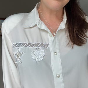 Puede incluir: Una camisa blanca abotonada con adornos de conchas marinas y estrellas de mar. La camisa presenta un collar de plata con colgante y pendientes a juego. La camisa tiene botones perlados y un ribete decorativo encima de los adornos.