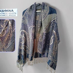 Puede incluir: Un chal Pashmina con un diseño de cachemira en tonos azules, grises y dorados. El chal está hecho de 70% cachemira y 30% seda. El chal tiene flecos en el borde inferior. El texto "Pashmina" es visible.
