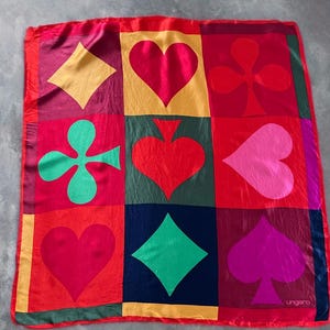Può includere: Una sciarpa quadrata di seta rossa con un motivo ripetuto di semi di carte da gioco in vari colori. I semi includono cuori, quadri, fiori e picche. La sciarpa ha un bordo rosso e il marchio "ungaro" è visibile.