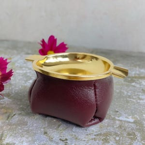 May include: A burgundy leather ashtray with a gold-colored interior and rim. The ashtray has a rounded shape and a small, built-in rest for a cigarette or cigar. The design is simple and elegant.