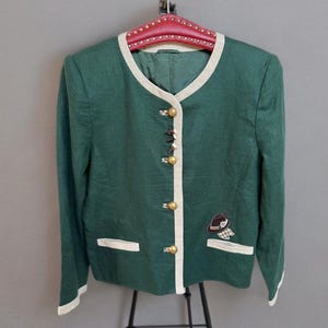 Puede incluir: Chaqueta verde con ribete crema y botones dorados. La chaqueta tiene un pequeño parche bordado con un diseño de sombrero y bufanda. Colgada en una percha roja.