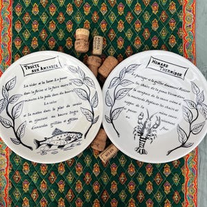 Puede incluir: Dos platos de cerámica blanca con texto negro e ilustraciones de un pez y una langosta. Los platos están sobre un mantel verde estampado con corchos esparcidos entre ellos. Los platos tienen texto en francés.