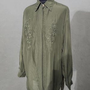 Puede incluir: Camisa verde oliva con botones, mangas largas y cuello clásico. La camisa presenta diseños florales bordados en la parte delantera. La tela tiene un ligero brillo, lo que sugiere seda o satén.