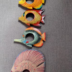 Puede incluir: Cuatro servilleteros de madera con forma de pez y colores vibrantes. Los peces están pintados en tonos amarillos, naranjas, rojos, azules y blancos. Cada uno tiene una abertura circular en el centro. Los servilleteros están dispuestos verticalmente sobre una superficie gris texturizada.
