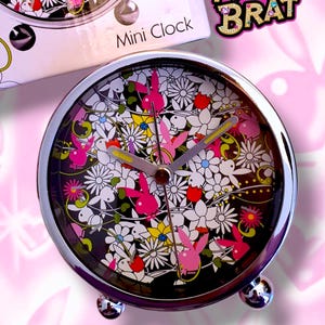 May include: A silver mini clock with a black face featuring a floral design with pink bunnies. The clock has a silver stand with three legs. The clock face also has the text "Pretty Brat" on it.