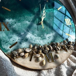 Puede incluir: Reloj de pared redondo con un diseño de océano de resina. La esfera del reloj presenta números romanos dorados, manecillas doradas y una escena de playa con conchas y pequeñas piedras. Los colores del reloj incluyen verde azulado, blanco y dorado.