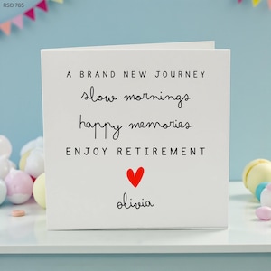 Peut inclure: Carte de retraite blanche avec texte noir : "Un tout nouveau voyage, des matins lents, de joyeux souvenirs, profitez de la retraite." Un cœur rouge et le nom "Olivia" sont également sur la carte. La carte est sur une surface blanche.