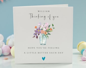 May include: A white greeting card with the text "William Thinking of you" and a watercolor floral arrangement in a glass vase. The card also includes the text "Hope you're feeling a little better each day" and a blue heart.