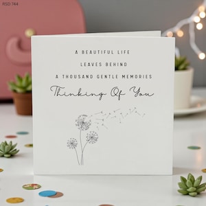 May include: A white greeting card with the message "A Beautiful Life Leaves Behind A Thousand Gentle Memories Thinking Of You." The card features a dandelion design. The card is on a white surface with decorative elements.