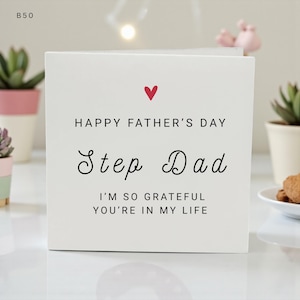 May include: A white Father's Day card with a red heart and the words "Happy Father's Day Step Dad". The message below reads "I'm so grateful you're in my life". The card is on a white surface.