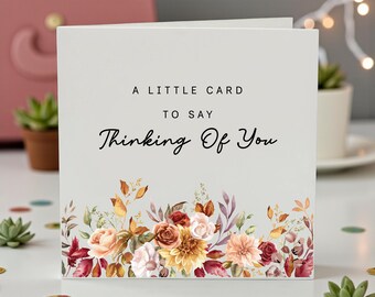 May include: A folded greeting card with the text "A Little Card To Say Thinking Of You." The card features a floral design with roses, dahlias, and other flowers in shades of peach, burgundy, and yellow. The card is on a white surface.