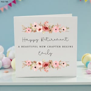 Op de afbeelding: Een witte pensioenkaart met bloemaccenten. De kaart bevat de tekst "Happy Retirement, A Beautiful New Chapter Begins, Emily" in elegant schrift. Roze en bordeauxrode bloemen sieren de boven- en onderkant.