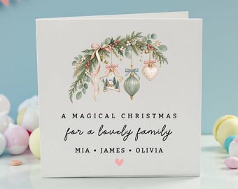 Pode incluir: Um cartão de felicitações quadrado branco com uma ilustração em aguarela de ramos de pinheiro decorados com ornamentos pendentes, incluindo uma estrela, uma gota e um coração. O texto diz: A MAGICAL CHRISTMAS for a lovely family MIA • JAMES • OLIVIA.