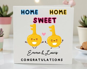Puede incluir: Una tarjeta blanca con las palabras "HOME SWEET HOME" en letras coloridas. Dos personajes de llaves amarillas con caras sonrientes y brazos aparecen debajo del texto. Los nombres "Emma & Lewis" y la palabra "CONGRATULATIONS" están impresos debajo.