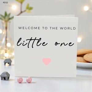 May include: A white greeting card with the text "WELCOME TO THE WORLD little one" in black script. A pink heart is below the text. Two pink candies and decorative objects are in the foreground.