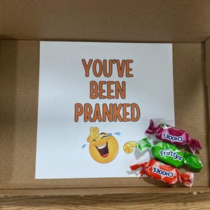 Op de afbeelding: Een witte kaart met de tekst "You've Been Pranked" en een cartoon huilende lachende emoji. Drie ingepakte snoepjes liggen voor de kaart. De snoepjes zijn rood, groen en oranje met het woord "Choose" erop gedrukt.