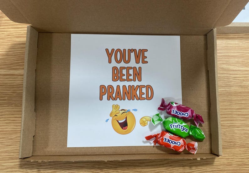 Op de afbeelding: Een witte kaart met de tekst "You've Been Pranked" en een cartoon huilende lachende emoji. Drie ingepakte snoepjes liggen voor de kaart. De snoepjes zijn rood, groen en oranje met het woord "Choose" erop gedrukt.