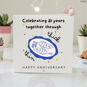 Könnte beinhalten: Weiße Jubiläumskarte mit dem Text "Celebrating 21 years together through thick & thin". Die Karte zeigt ein blaues Oval mit zwei Fischillustrationen und den Worten "Happy Anniversary".